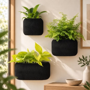 Peut inclure: Trois jardinières murales noires avec un motif ondulé vertical texturé. Chaque jardinière contient une plante verte. Les jardinières sont disposées sur un mur, avec une jardinière carrée plus petite au-dessus et deux rectangulaires en dessous.