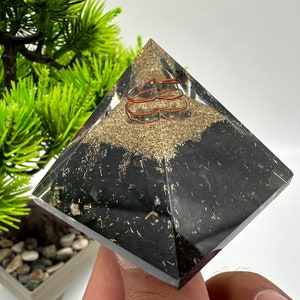 May include: A clear crystal pyramid with a black base and a copper wire coil inside. The pyramid is filled with black and gold glitter.