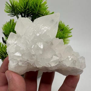 Peut inclure: Un amas de cristaux de quartz blancs, avec une texture rugueuse et naturelle. Les cristaux sont disposés en grappe, certains pointant vers le haut.
