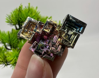 May include: A colorful bismuth crystal with a geometric, stepped structure. The crystal displays iridescent hues of gold, purple, blue, and green. The unique mineral specimen is held against a blurred green background.