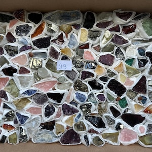 Op de afbeelding: Een verzameling van talrijke ruwe mineraalspecimens, elk afzonderlijk verpakt in wit papier en strak gerangschikt in een rechthoekige kartonnen doos. Een klein wit vierkant label met het handgeschreven getal 99 ligt in het midden.