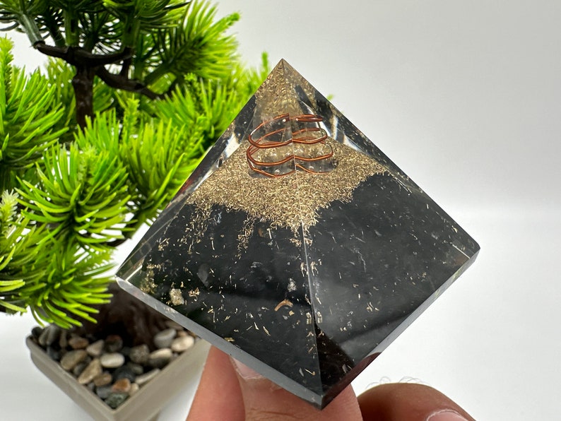 May include: A clear crystal pyramid with a black obsidian base and a copper wire coil inside. The pyramid is sitting on a hand.