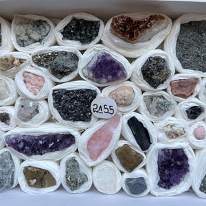 Puede incluir: Una colección de varios especímenes minerales en bruto y cristales, cada uno envuelto individualmente en papel de seda blanco y dispuesto en una caja. Una pequeña etiqueta circular blanca con el número 2155 escrito a mano se coloca entre las piedras, que incluyen amatista morada, cuarzo rosa, turmalina negra y pirita metálica.