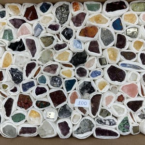 Könnte beinhalten: Eine Sammlung von 100 verschiedenen rohen Mineralproben und Kristallen, die jeweils einzeln in weißes Papier eingewickelt und in einem Raster in einem Karton angeordnet sind. Die Steine zeigen eine Vielzahl von Farben, darunter Violett, Gelb, Grün und Schwarz.