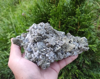 May include: A large, rough, gray and gold rock with a crystalline texture. The rock is being held in a hand.