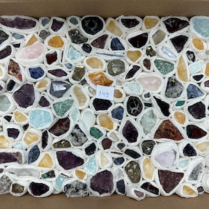 Könnte beinhalten: Eine Sammlung verschiedener roher Mineralsteine in verschiedenen Farben, darunter Violett, Gelb, Blau und Transparent, die jeweils einzeln in weißes Papier eingewickelt und in einem Karton angeordnet sind. Ein kleines weißes Etikett mit der Nummer 149 ist sichtbar.