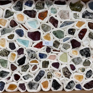Könnte beinhalten: Eine Sammlung verschiedener roher Mineralexemplare, die jeweils einzeln in weißen Stoff eingewickelt und in einem Raster in einer Schachtel angeordnet sind. Die Steine zeigen verschiedene Farben wie Violett, Gelb, Blau, Grün und Schwarz. Ein kleines weißes Etikett mit der Aufschrift 45 ist sichtbar.