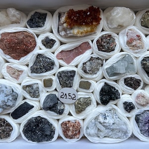 Könnte beinhalten: Eine Sammlung verschiedener roher Mineralproben, die einzeln in weißes Seidenpapier eingewickelt und in einem Karton angeordnet sind. Das Sortiment umfasst Steine in Weiß-, Grau-, Schwarz-, Braun-, Orange- und Violetttönen sowie ein weißes Etikett mit der Aufschrift 2152.