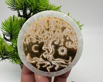 May include: A white round coaster with a gold tree of life design. The tree has intricate branches and leaves, and there is a crescent moon and a sun on either side of the trunk.