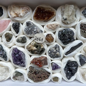 Könnte beinhalten: Eine Sammlung verschiedener roher Mineralproben und Kristalle, die jeweils einzeln in weißes Seidenpapier eingewickelt und in einer rechteckigen Schachtel angeordnet sind. Ein kleines rundes weißes Etikett mit der handgeschriebenen Nummer 2161 befindet sich in der Mitte.