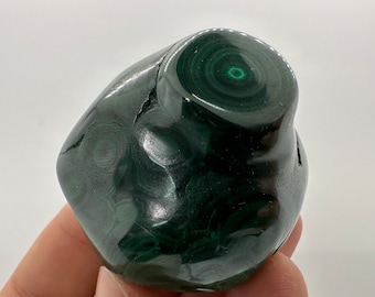 May include: A polished green malachite stone with a swirling pattern. The stone is smooth and has a natural, organic shape.