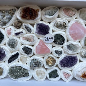 Op de afbeelding: Een verzameling van diverse ruwe mineralen, elk afzonderlijk verpakt in wit papier en gerangschikt in een rechthoekige doos. De stenen omvatten paarse amethist, roze rozenkwarts, metaalachtige pyriet en andere kristallen. Een wit label toont het getal 2154.