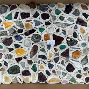 Könnte beinhalten: Eine Sammlung verschiedener roher Mineralsteine und Kristalle, die jeweils einzeln in weißes Papier eingewickelt und dicht in einem rechteckigen Karton angeordnet sind. Ein kleines weißes Etikett mit der handschriftlichen Zahl 119 liegt mittig.