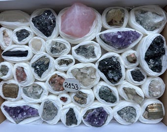 May include: A collection of various raw mineral specimens individually wrapped in white tissue paper and arranged in a box. The assortment includes a large pink rose quartz, purple amethyst clusters, metallic pyrite, and dark grey ore samples. A small white circular tag with the handwritten number 2153 is placed among the stones.