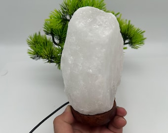 Op de afbeelding: Een witte Himalaya zoutlamp met een ruwe, natuurlijke textuur. De lamp staat op een ronde, donkere houten voet en heeft een zwart netsnoer. Groene decoratieve bladeren zijn op de achtergrond.