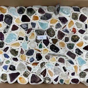 Könnte beinhalten: Eine Sammlung verschiedener roher Mineralproben und Kristalle, die jeweils einzeln in weißes Papier eingewickelt und in einem rechteckigen Karton angeordnet sind. Ein kleines weißes Etikett mit dem handschriftlichen Text 69 befindet sich in der Mitte.