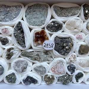 Könnte beinhalten: Eine Sammlung verschiedener roher Mineralexemplare, die einzeln in weißes Seidenpapier eingewickelt und in einer rechteckigen Schachtel angeordnet sind. Ein Exemplar trägt ein weißes, rundes Etikett mit der handschriftlichen Nummer 2158. Die Steine variieren in Farbe, Textur und Kristallstruktur, einschließlich metallischer, grauer, weißer und bernsteinfarbener Töne.