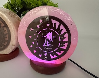 Op de afbeelding: Een roze, ronde zoutlamp met een zwart Maagd dierenriem ontwerp. De lamp staat op een houten voet en straalt een zachte, kleurrijke gloed uit. Het ontwerp bevat sterren, zonnestralen en het Maagd-symbool.