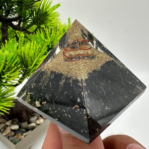 May include: A black crystal pyramid with a copper wire coil inside. The pyramid is sitting on a hand.