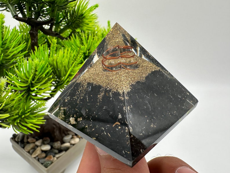 May include: A black crystal pyramid with a copper wire coil inside. The pyramid is sitting on a hand.