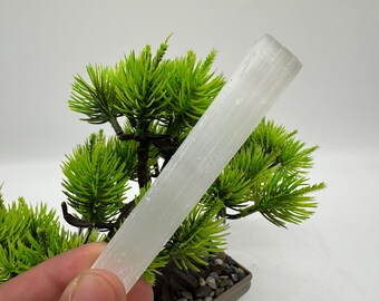 May include: A white selenite wand, also known as a cleansing wand, is held in a hand in front of a small artificial bonsai tree. The wand is smooth and has a striated texture.
