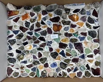 May include: A collection of assorted raw mineral specimens and crystal clusters individually wrapped in white paper, arranged in a rectangular cardboard box. A small white paper tag with the handwritten number 127 is placed in the centre.