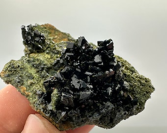 May include: A cluster of black crystals on a green rock. The crystals are sharp and have a metallic sheen. The rock is rough and textured.