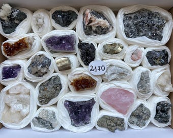 Può includere: Una collezione di vari esemplari di minerali grezzi e grappoli di cristalli avvolti singolarmente in carta velina bianca e disposti in una scatola. L'assortimento comprende ametista viola, pirite dorata, quarzo rosa e minerali grigio scuro con un'etichetta bianca contrassegnata con 2170.