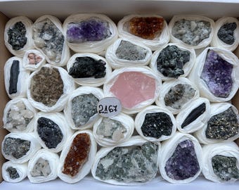 May include: A collection of various raw mineral specimens and crystal clusters individually wrapped in white paper, arranged in a box. The assortment includes purple amethyst, pink rose quartz, dark pyrite, and black tourmaline, with a tag labelled 2167.
