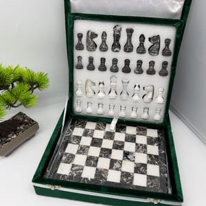 Könnte beinhalten: Ein Schachspiel mit einem Marmorbrett mit abwechselnd schwarzen und weißen Feldern, untergebracht in einem grün mit Samt ausgekleideten Koffer. Das Set enthält eine vollständige Sammlung schwarzer und weißer Marmor-Schachfiguren in einer Schaumstoffeinlage.