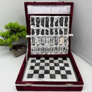 Könnte beinhalten: Ein Schachspiel mit einem schwarz-weißen Marmorbrett und passenden geschnitzten Steinschachfiguren. Die Figuren sind in einem mit Samt ausgekleideten Koffer mit weißem Satin-Innenfutter untergebracht. Der Koffer ist aus dunkelrotem Samt.