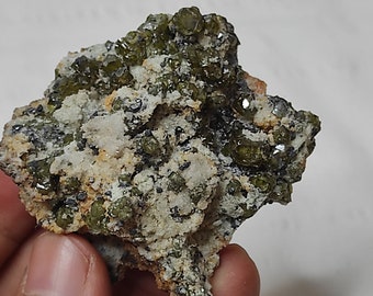May include: A rough, gray rock with green crystals embedded in it. The rock has a white, powdery coating in some areas.