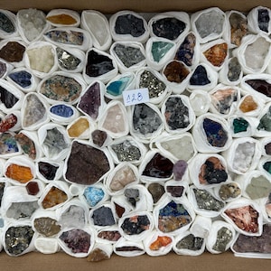 Könnte beinhalten: Eine Sammlung verschiedener roher Mineralproben und Kristalle, die jeweils einzeln in weißes Papier eingewickelt und in einem Karton angeordnet sind. Ein kleines weißes Etikett mit der handschriftlichen Nummer 128 befindet sich in der Mitte.