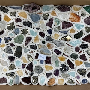 Könnte beinhalten: Eine Sammlung verschiedener roher Mineralien und Kristalle, die jeweils einzeln in weißes Papier eingewickelt und in einem rechteckigen Karton angeordnet sind. Ein kleines weißes Etikett mit der handschriftlichen Zahl 153 liegt in der Mitte.