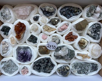 Può includere: Una collezione di vari esemplari di minerali grezzi e cristalli, ciascuno avvolto singolarmente in carta velina bianca e disposto in una scatola. Una piccola etichetta circolare bianca con il numero 2159 scritto a mano è posizionata tra le pietre.