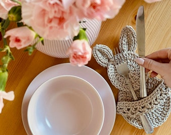 May include: A crocheted bunny-shaped utensil holder with a fork and knife inside. The holder is beige and the utensils are silver. The holder is on a wooden table next to a pink bowl.