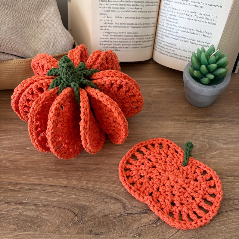 May include: A three-dimensional crocheted orange pumpkin with a dark green stem and a matching flat, circular orange crocheted coaster with a small green stem, both featuring an open-work stitch pattern on a wooden surface.