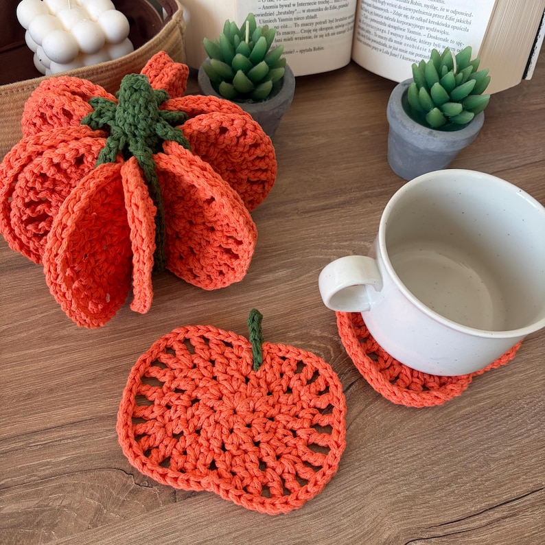 May include: A set of orange crochet pumpkin coasters, with one coaster placed under a white ceramic mug and another flat on the surface. The coasters feature a textured, open-stitch design with a small green stem at the top, mimicking the shape of a pumpkin.
