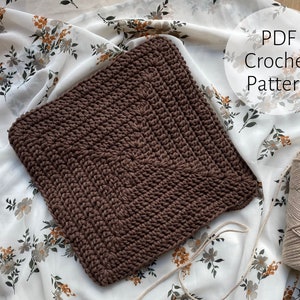 Puede incluir: Un artículo cuadrado de ganchillo marrón, posiblemente un salvamanteles o un paño, se muestra sobre una tela con estampado floral. El texto "PDF Crochet Pattern" es visible. Una madeja de hilo beige está en la esquina inferior derecha.