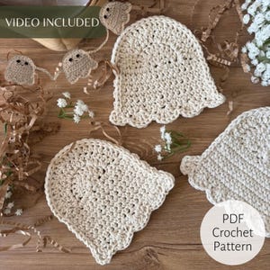May include: Three off-white crochet ghost shapes with scalloped edges and two small black circular eyes each. The ghosts are arranged on a wooden surface alongside brown paper shreds and small white flowers. Text reads VIDEO INCLUDED and PDF Crochet Pattern.