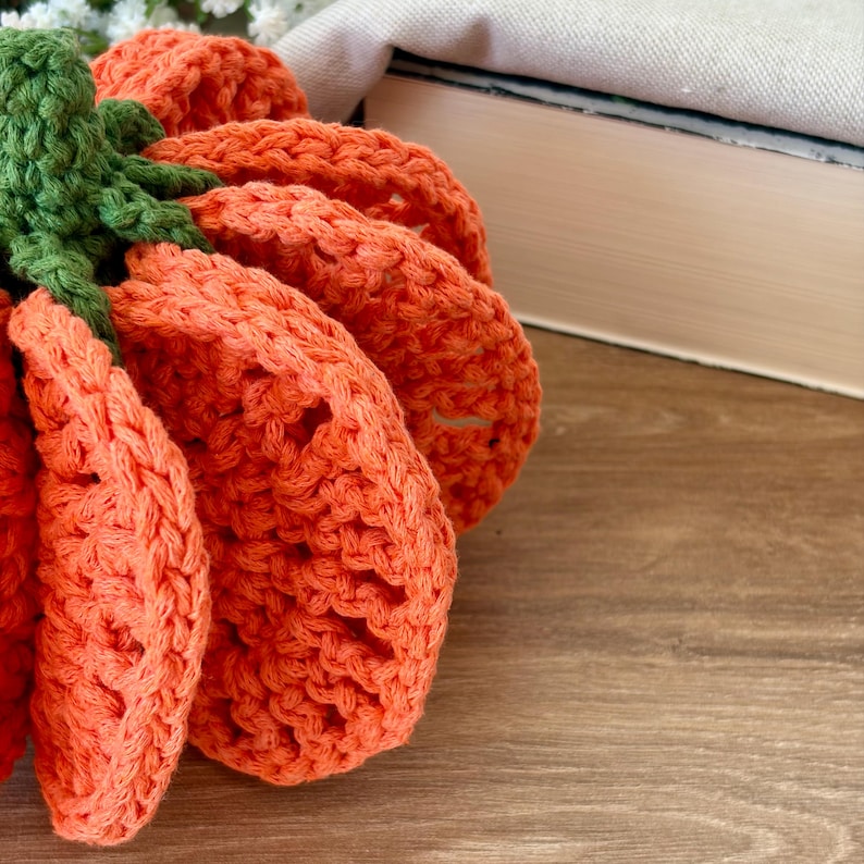 May include: A handcrafted crochet pumpkin featuring a vibrant orange body with a textured, ribbed stitch pattern and a solid green stem at the top. The pumpkin is constructed from yarn, showcasing a rounded, segmented shape with visible loops.