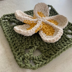 May include: A crocheted green square with a white and yellow crocheted flower in the center. The flower has five petals and is made with a yellow and white yarn.