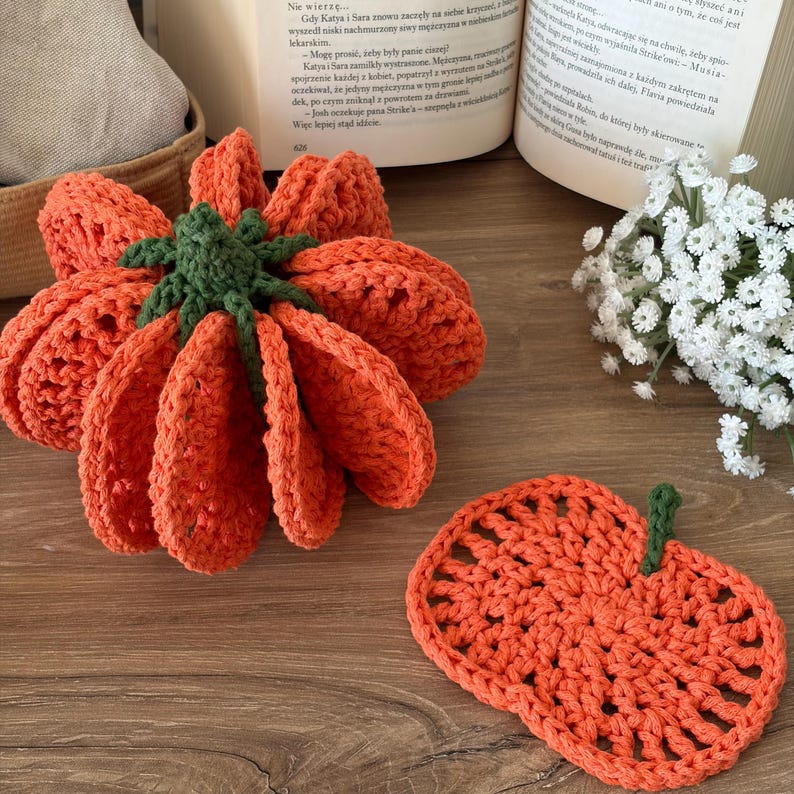 May include: A three-dimensional crocheted orange pumpkin with a dark green stem and a separate flat, circular crocheted orange coaster featuring a small green stem, both crafted from yarn with visible open-stitch patterns and textured surfaces.