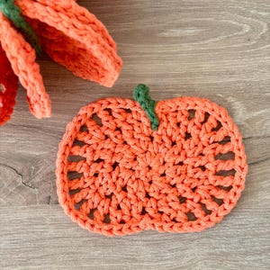 May include: A flat, orange, pumpkin-shaped crochet coaster with an openwork stitch pattern and a small, dark green crocheted stem at the top. A portion of a larger, three-dimensional orange crochet pumpkin is visible in the upper left corner.