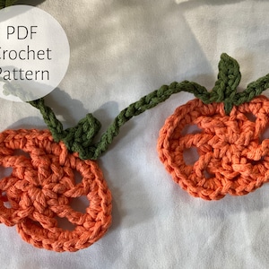 Può includere: Una ghirlanda all'uncinetto con zucche arancioni e foglie verdi. La ghirlanda è realizzata con un punto semplice e ha un aspetto rustico e fatto a mano. Il testo "PDF Crochet Pattern" è visibile nell'immagine.