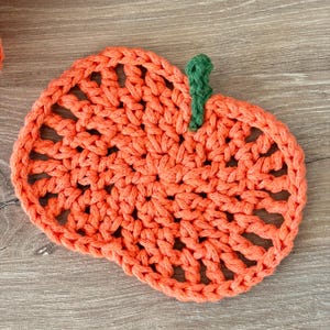 May include: A round, orange crochet coaster shaped like a pumpkin, featuring an openwork stitch pattern and a small, textured green stem attached to the top edge. The yarn is thick and creates a visible, repeating loop design throughout the piece.