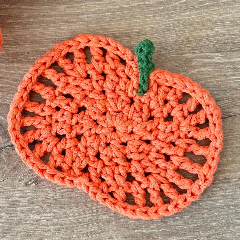 May include: A round, orange crochet coaster shaped like a pumpkin, featuring an openwork stitch pattern and a small, textured green stem attached to the top edge. The yarn is thick and creates a visible, repeating loop design throughout the piece.