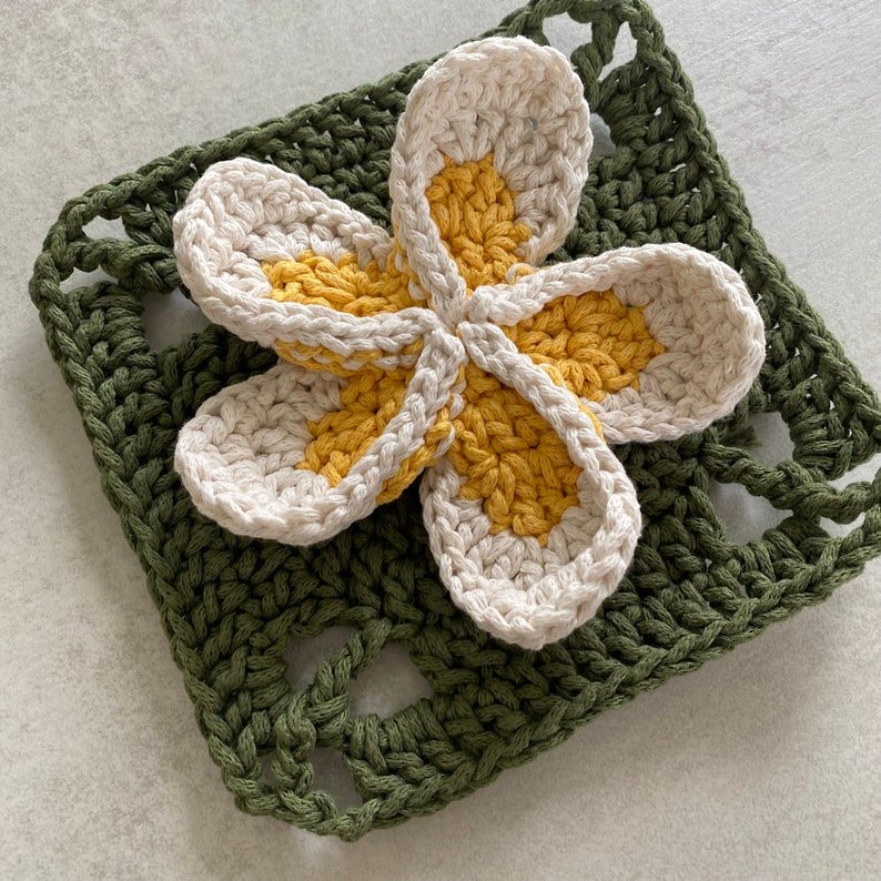 May include: A crocheted flower with white petals and yellow centers is placed on a green crocheted square.