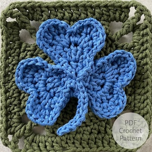 Könnte beinhalten: Ein blaues gehäkeltes vierblättriges Kleeblatt-Muster auf einem grünen gehäkelten quadratischen Hintergrund. Der Text "PDF Crochet Pattern" befindet sich in der unteren rechten Ecke des Bildes.