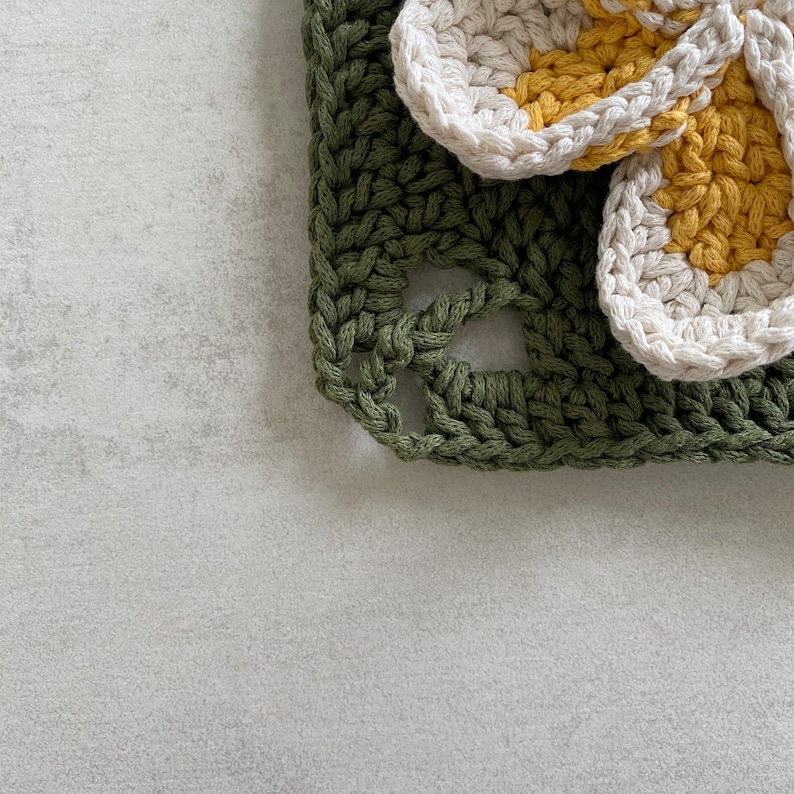 May include: A crocheted green square with two white and yellow crocheted flowers on top.